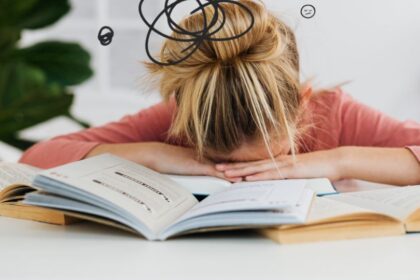 Egy fiatal nő könyvek között ül, kezét a fejére téve, a halogatás és stressz jeleit mutatja.