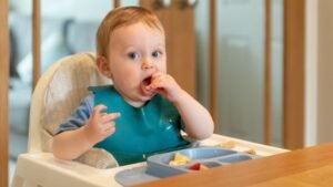 Egy kisgyermek ül egy etetőszékben, ételt eszik, miközben figyelmesen néz.
