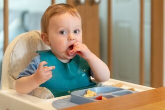 Egy kisgyermek ül egy etetőszékben, ételt eszik, miközben figyelmesen néz.