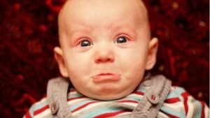 Egy kisbaba szomorúan néz, könnycsepp csillog a szemében, a háttér piros szőnyeg.