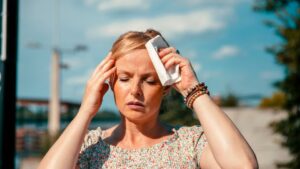 womensway 111 Egy nő, aki a fejét fogja, láthatóan napszúrástól szenved, törölközőt használva a hűtéshez.