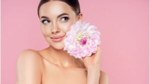 Egy fiatal nő mosolyogva tart egy nagy virágot az arcához, háttérben rózsaszín fal.