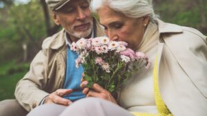 womensway 117 Idős pár ül egy parkban, a nő virágcsokrot szagol, míg a férfi figyeli őt.