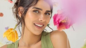 Egy fiatal nő mosolyog, zöld topban, virágokkal körülvéve, ragyogó bőrrel.