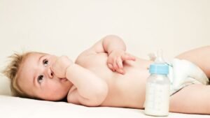Egy kisbaba fekszik a hátán, ujja a szájában, mellette egy cumisüveg.