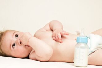 Egy kisbaba fekszik a hátán, ujja a szájában, mellette egy cumisüveg.