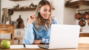 Egy fiatal nő mosolyogva ül egy asztalnál, laptop előtt, miközben egy ceruzát tart a kezében.