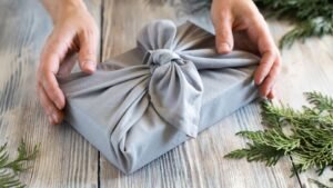 womensway 145 Egy kéz tart egy szürke ajándékcsomagot, amelyet szép csomagolás díszít, fenyőágakkal körülvéve.