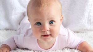 Egy mosolygó baba hason fekszik egy puha, fehér szőnyegen, élénk kék szemekkel.