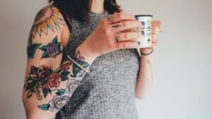 womensway 135 Egy nő, aki tetoválásokkal díszített karját mutatja, miközben egy bögrét tart a kezében.