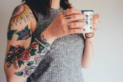 Egy nő, aki tetoválásokkal díszített karját mutatja, miközben egy bögrét tart a kezében.