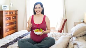 Egy várandós nő ül a ágyon, salátát eszik, mosolyogva néz a kamerába.
