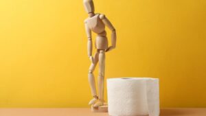 womensway 124 Egy fából készült figura, amely a vécépapír mellett áll, sárga háttér előtt.