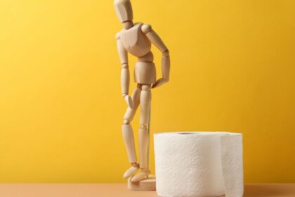 Egy fából készült figura, amely a vécépapír mellett áll, sárga háttér előtt.