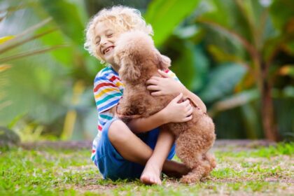 Egy boldog kisfiú öleli a kutyáját a zöld fűben, mosolyogva játszanak.