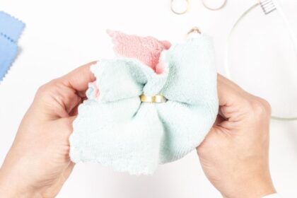 Arany ékszerek otthoni tisztítása – mutatunk 4 praktikus tippet 2 Két kéz egy arany gyűrűt tisztít egy színes mikroszálas kendőben, háttérben tisztító eszközök.