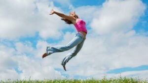 Egy fiatal nő boldogan ugrik a zöld fűben, a kék ég alatt, kifejezve a szabadság érzését.