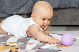 Az első mosolytól az első lépésekig: a baba fejlődésének titkos naptára 3 Egy kisbaba hason fekszik a szőnyegen, és egy lila labdát próbál elérni.