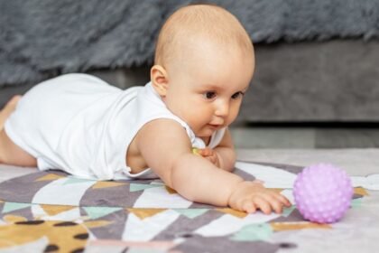 Egy kisbaba hason fekszik a szőnyegen, és egy lila labdát próbál elérni.