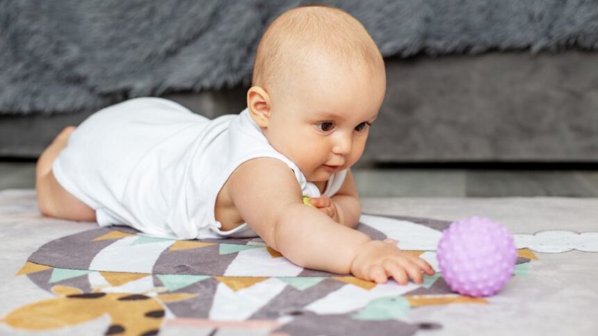 Egy kisbaba hason fekszik a szőnyegen, és egy lila labdát próbál elérni.