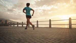 womensway 128 Egy fiatal nő fut a tengerparti sétányon, napfelkeltében, energikus mozdulattal.