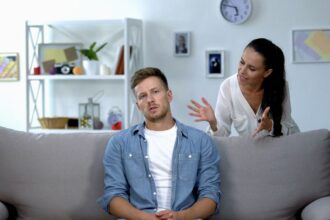 Egy flegmatikus férfi ül a kanapén, míg egy nő beszél hozzá, láthatóan érzelmi helyzetben.