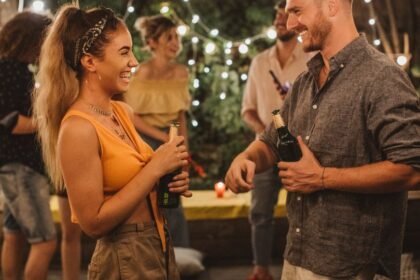 Egy fiatal pár nevetve beszélget egy baráti összejövetelen, italokkal a kezükben.