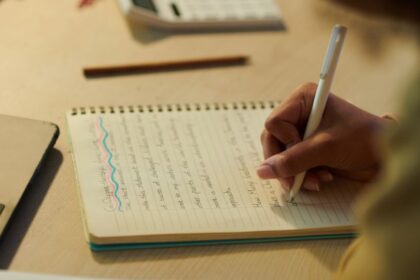 Egy kéz ír egy jegyzetfüzetbe, mellette laptop és tollak találhatók, ami a vázlatírás folyamatát ábrázolja.