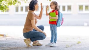 womensway 141 Egy anya és lánya vidáman integetnek egymásnak az iskola előtt, a lány iskolatáskával és füzetekkel a kezében.