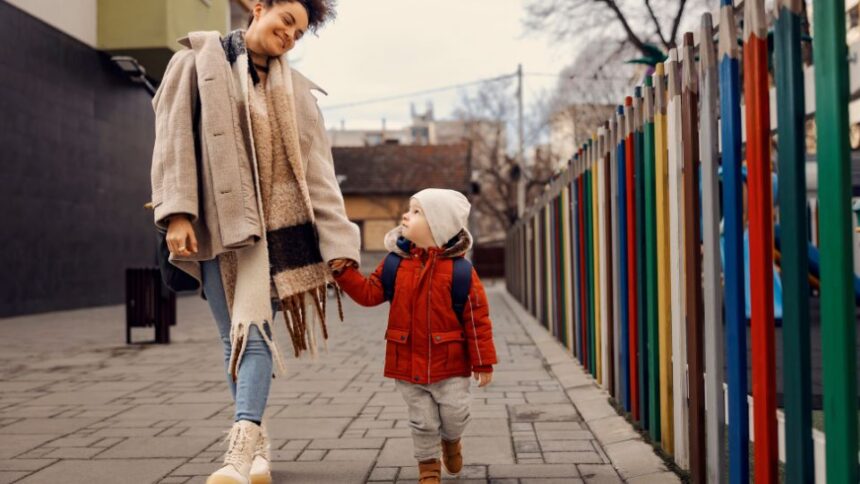 Hogyan készítsük fel a gyerekünket az óvodára? 1 Egy anya és kisgyermeke kézen fogva sétálnak egy színes kerítés mellett, az óvodába tartva.
