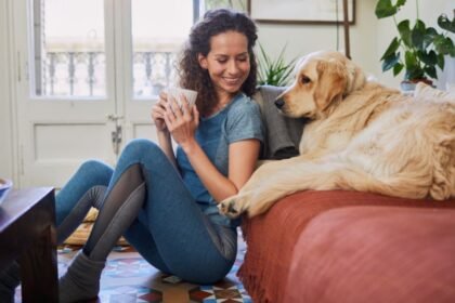 Egy fiatal nő ül a padlón, kezében egy csésze, mellette egy arany retriever pihen.