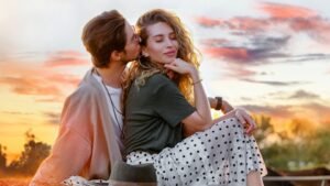 Egy fiatal pár ül egy gyönyörű naplementében, a férfi a nő füléhez hajol, miközben a nő mosolyogva néz előre.