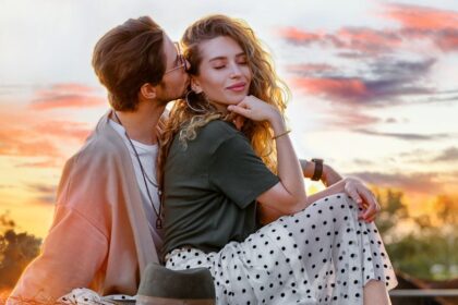 Egy fiatal pár ül egy gyönyörű naplementében, a férfi a nő füléhez hajol, miközben a nő mosolyogva néz előre.