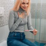 Egy fiatal nő ül a fürdőszobában, kezében terhességi teszt, arca boldog és izgatott.