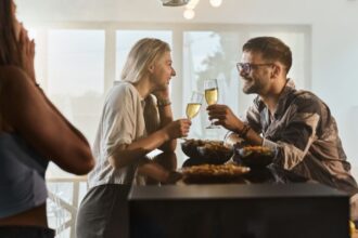 Egy férfi és egy nő pezsgőt koccintanak egy baráti összejövetelen, miközben egy harmadik személy figyeli őket.