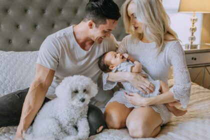 Egy fiatal pár boldogan ül az ágyon, a nő a kisbabáját tartja, míg a férfi mosolyogva nézi őket. Mellette egy fehér kutya ül.