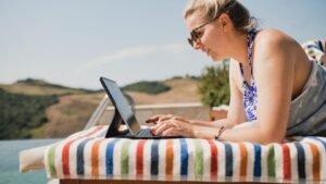 Egy nő napernyő alatt, laptopon dolgozik egy színes csíkos törölközőn, háttérben dombos táj.