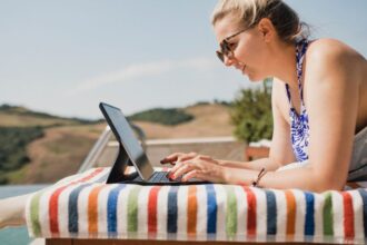 Egy nő napernyő alatt, laptopon dolgozik egy színes csíkos törölközőn, háttérben dombos táj.