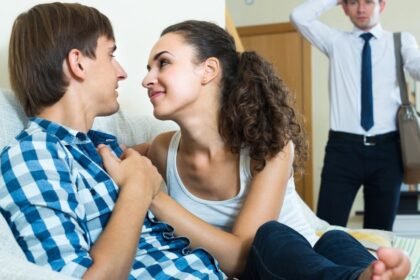 Egy fiatal pár közel ül egymáshoz a kanapén, miközben egy férfi háttérben, meglepetten nézi őket.