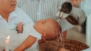 womensway 127 Egy kisbaba keresztelője, ahol víz ömlik a fejére, miközben a szülők és a pap jelen vannak.