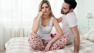 Egy nő ül a ágyon, gondterhelt arccal, míg a férfi mögötte próbálja megnyugtatni.