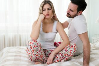 Egy nő ül a ágyon, gondterhelt arccal, míg a férfi mögötte próbálja megnyugtatni.