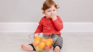 Egy kisbaba ül a földön, kezében gyümölcsöt tartva, egy tál gyümölcs előtt.