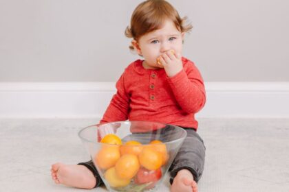 Egy kisbaba ül a földön, kezében gyümölcsöt tartva, egy tál gyümölcs előtt.