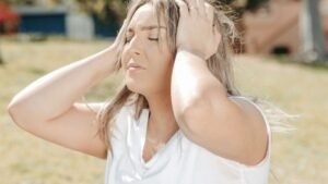 womensway 147 Egy fiatal nő a fejét fogja, láthatóan szorong vagy stresszes, a háttérben zöld fű és épületek.