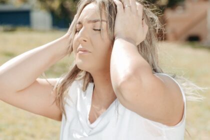 Egy fiatal nő a fejét fogja, láthatóan szorong vagy stresszes, a háttérben zöld fű és épületek.