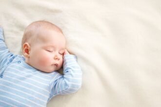 Egy kisbaba békésen alszik egy puha, világos takarón, kék csíkos ruhában.