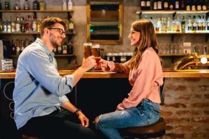 Két fiatal, mosolygós ember söröket koccint egy barátságos bárban, kellemes hangulatban.