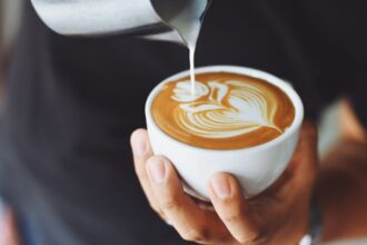 Egy kéz egy fehér csészében kávét önt, a kávé tetején szép latte art látható.
