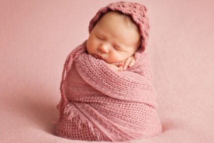 Egy újszülött csecsemő rózsaszín takaróba burkolva, kis sapkában, békésen alszik.